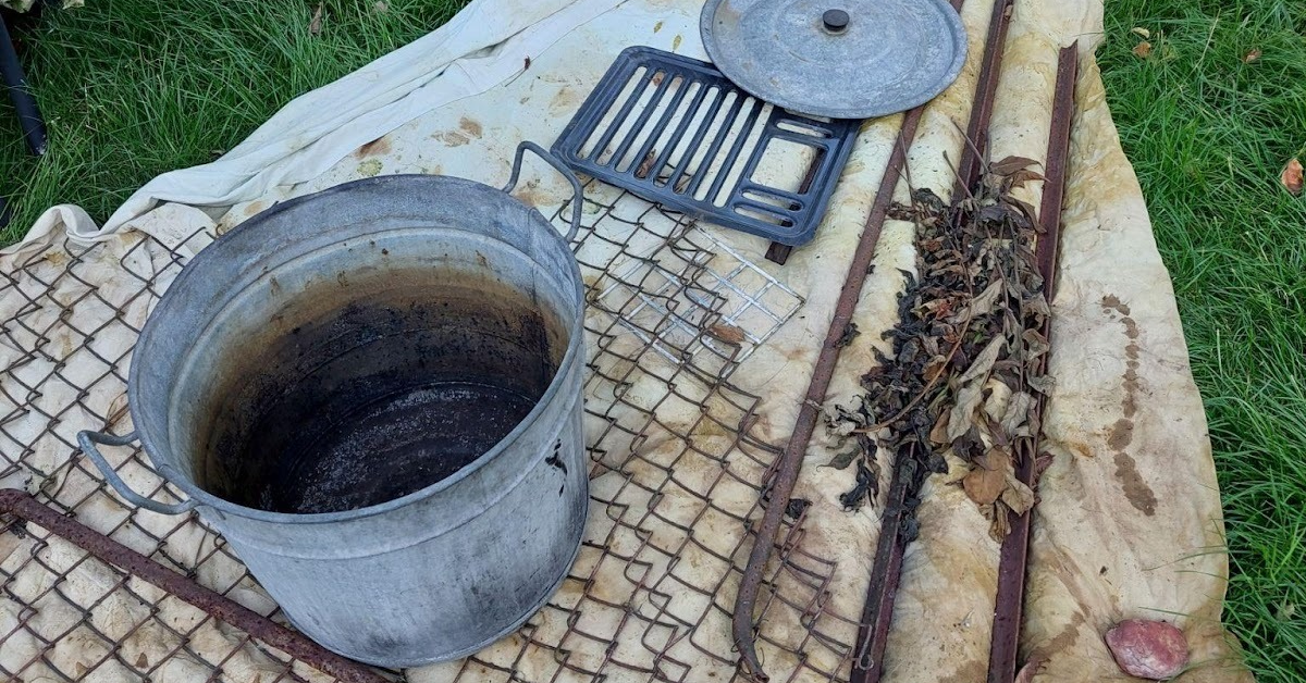 Panele tkaniny rozłożone na trawie w ogrodzie, dociśnięte rdzewiejącymi metalowymi przedmiotami, takimi jak garnek i siatka, w celu uzyskania śladów rdzy.